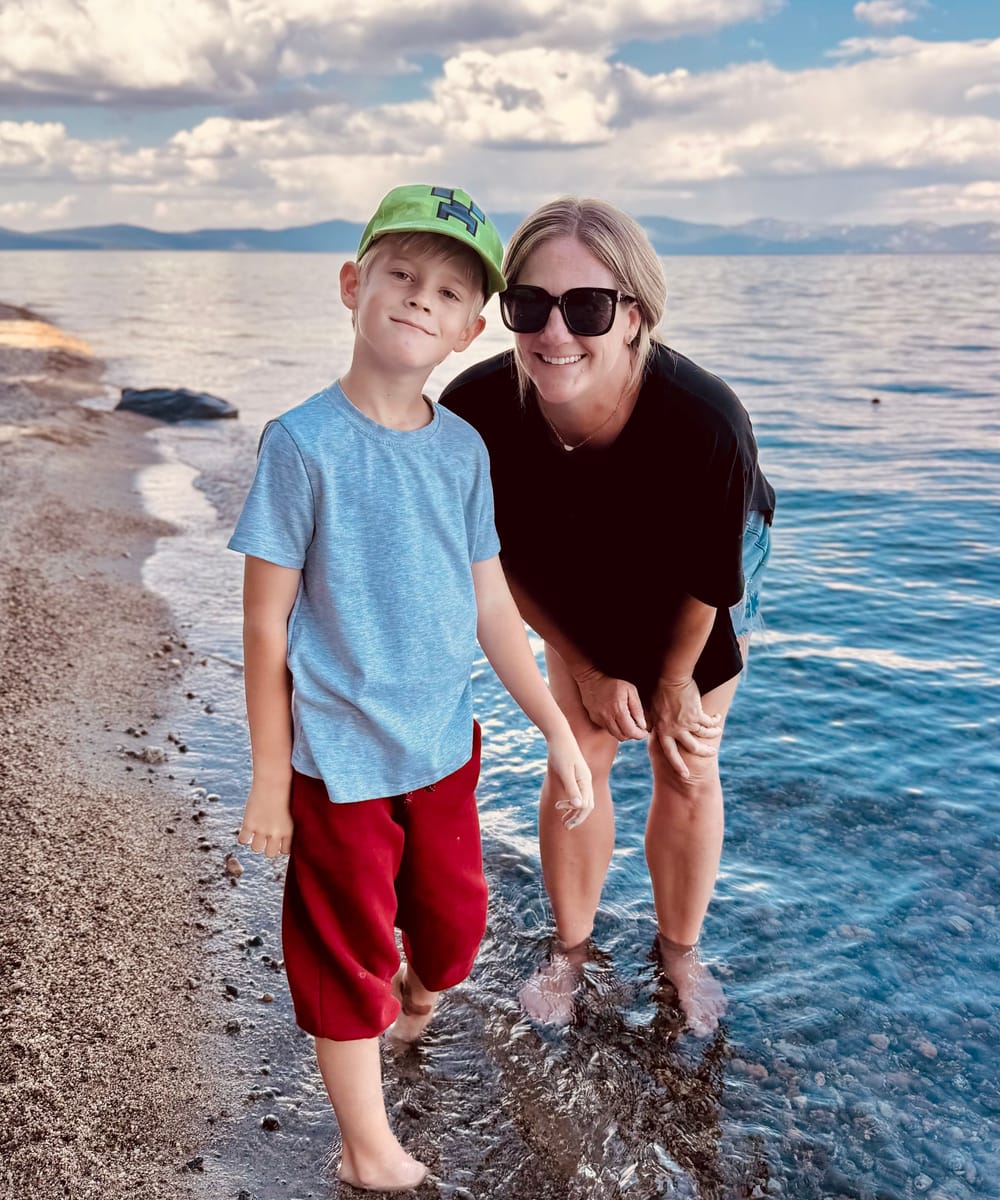 Samantha smiling beside a young boy at the edge of a lake, standing barefoot in the shallow water